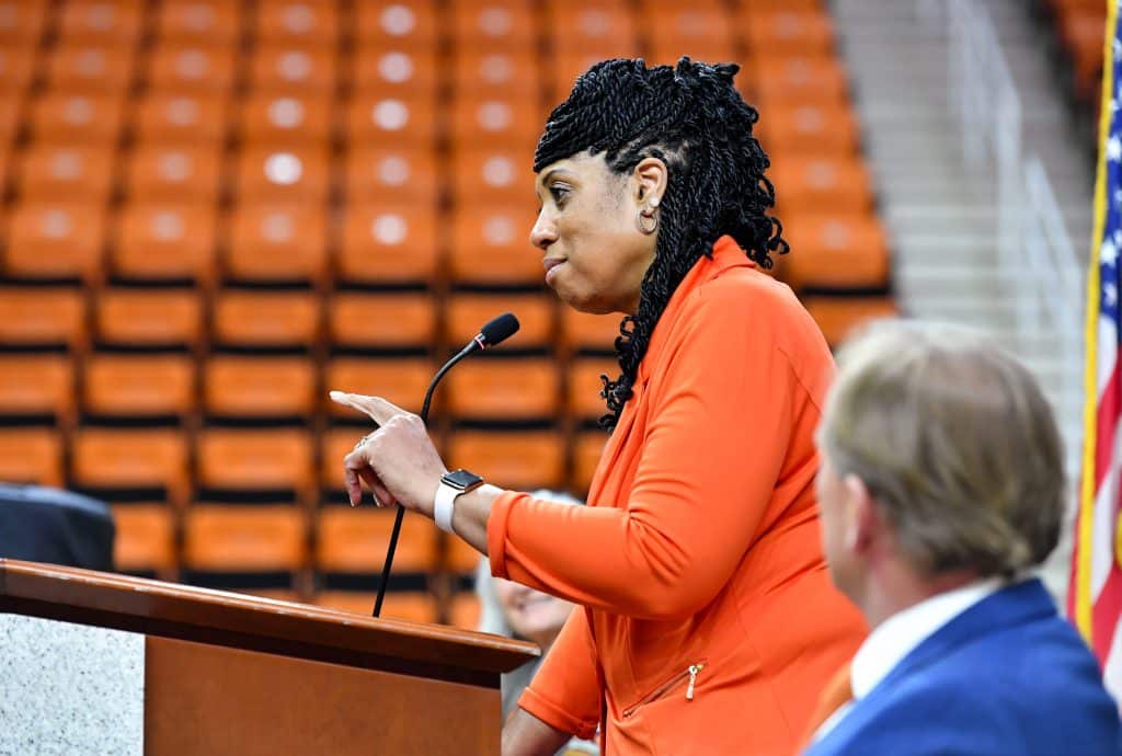 Michelle Clark-Heard, dressed all in orange, talks into a microphone while pointing her right hand.