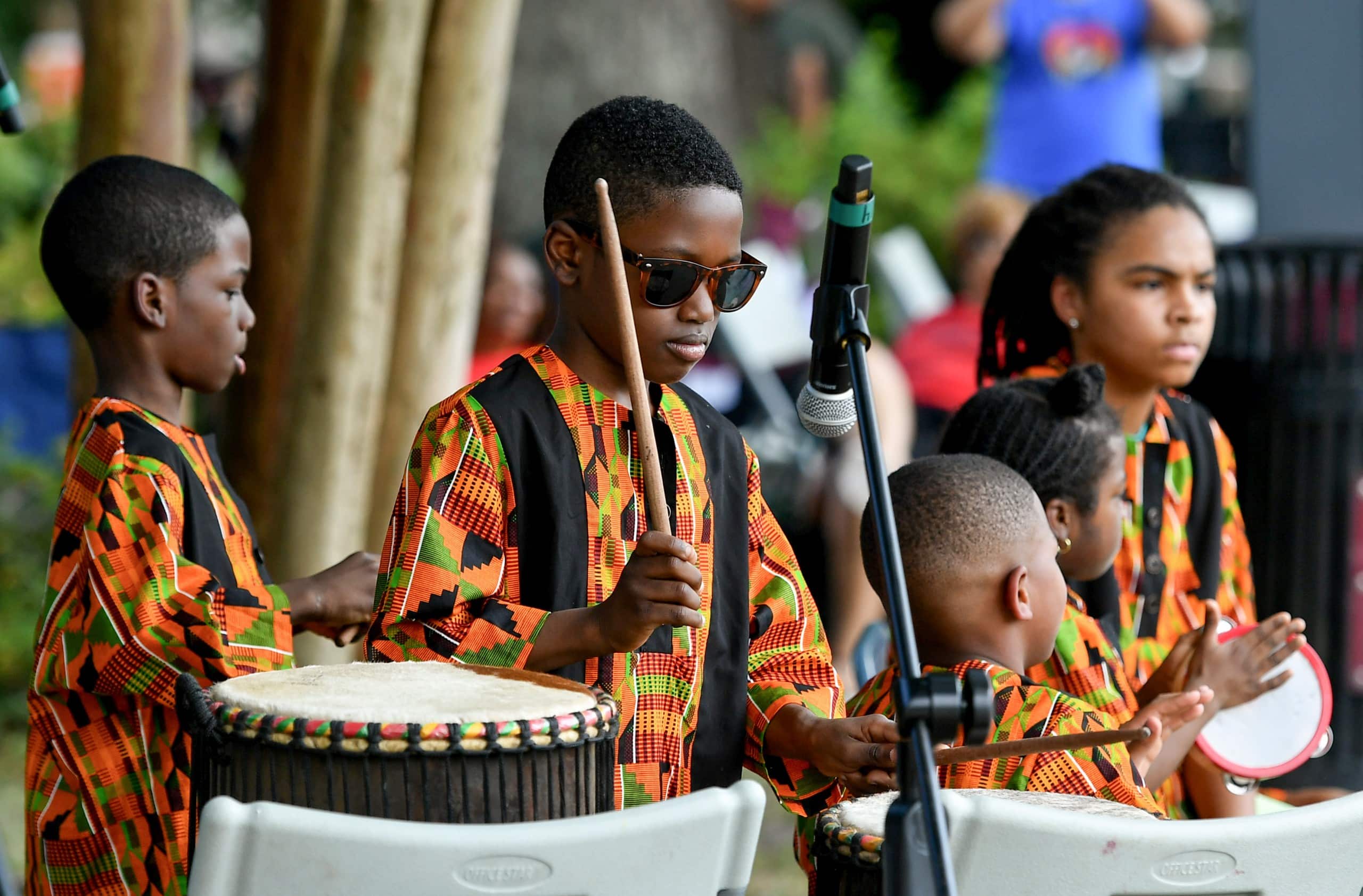 Several youth in colorful attire play drums in front of a microphone at a park.