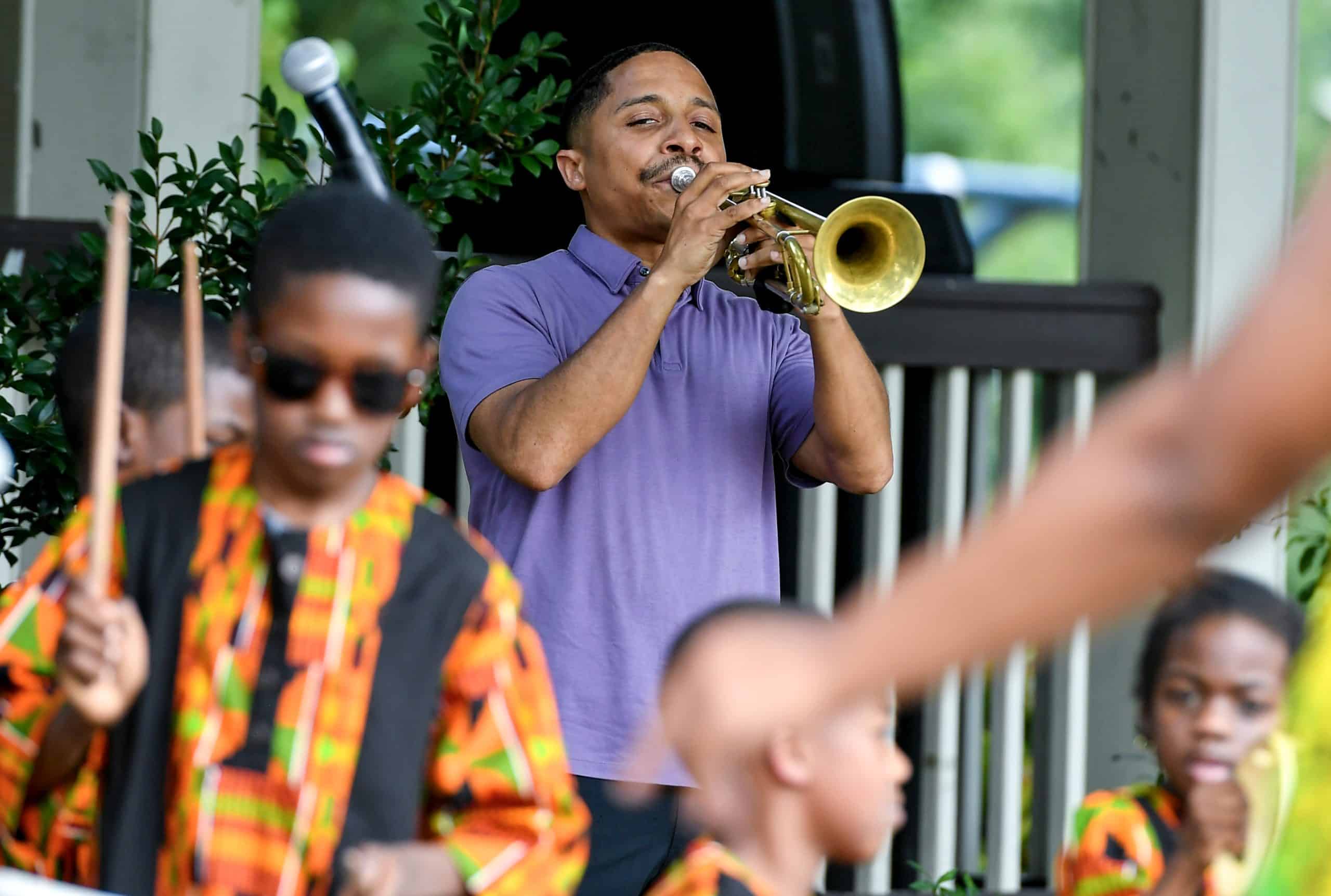 A man plays a trumpet as youth play drums in a park.