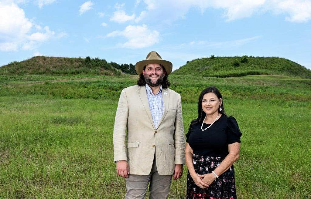 A man and a woman stand outside in front of two mounds covered in grass.