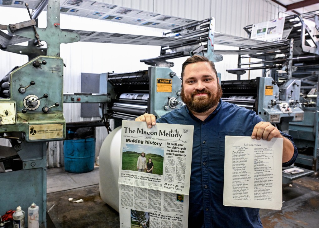 A man holds a newspaper and a newsletter printed on computer paper with press equipment in the background.