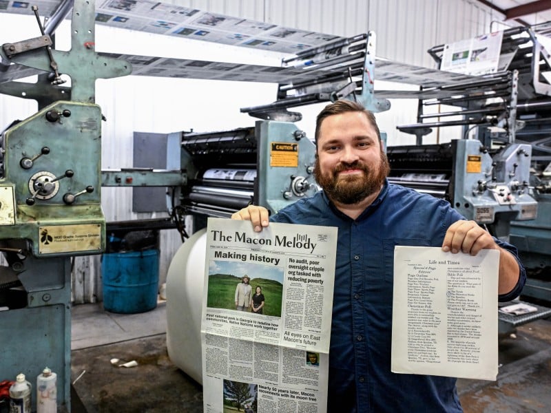 A man holds a newspaper and a newsletter printed on computer paper with press equipment in the background.