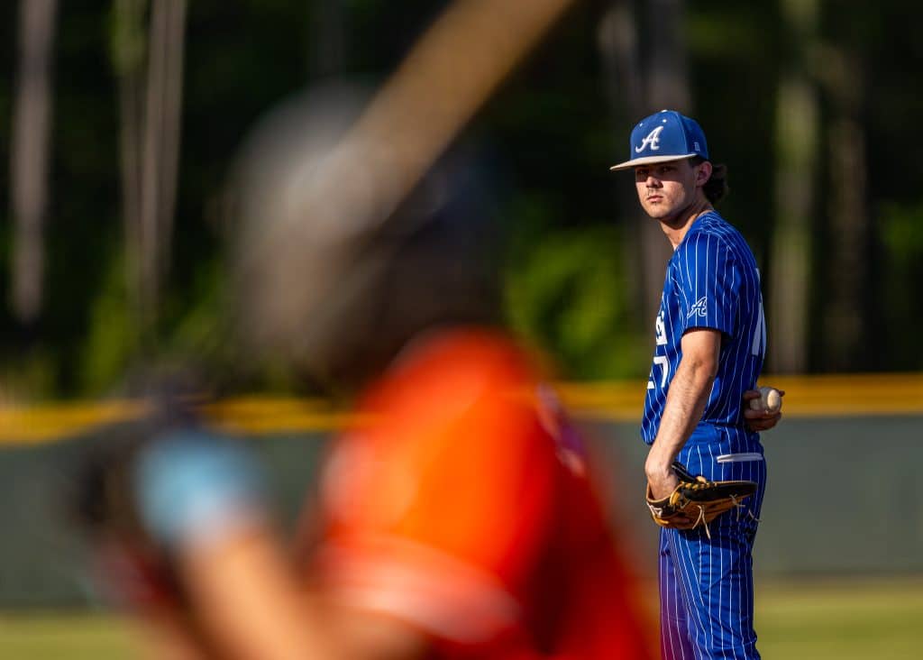 An ACE pitcher in a blue pinstriped uniform stands on the mound holding the baseball behind his back. An out-of-focus batter stands in the foreground.
