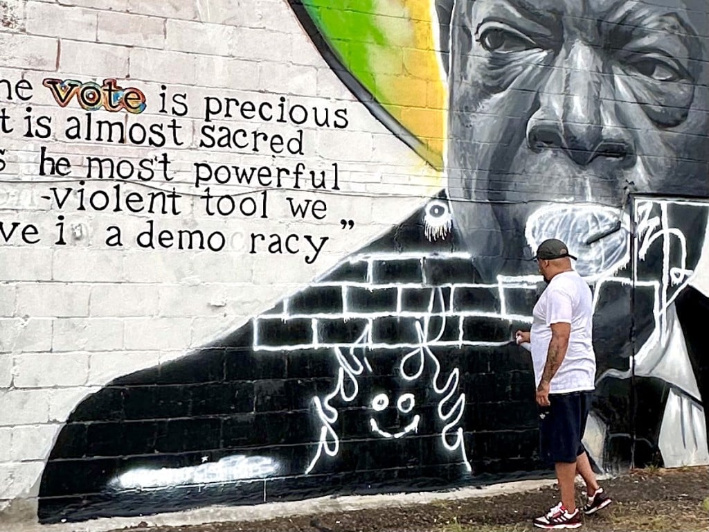 An artist holds a spray can in hand in front of a mural on a brick wall.