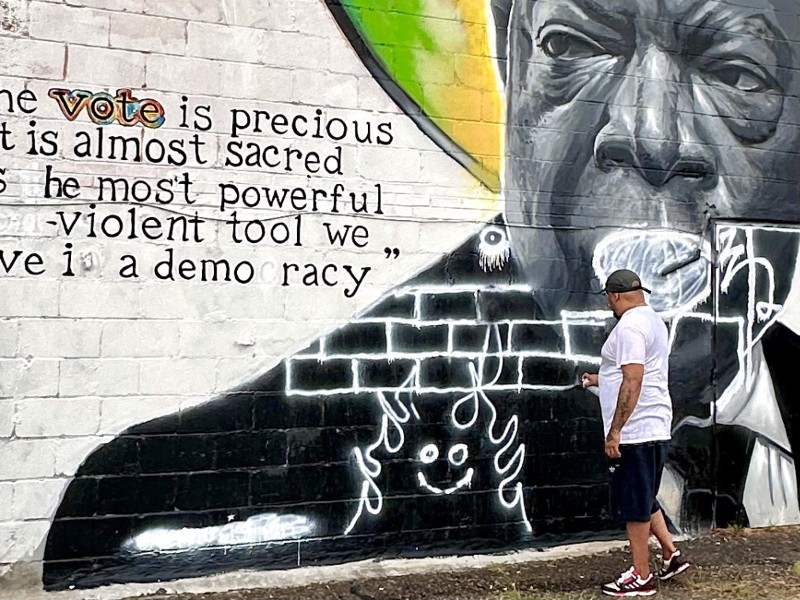 An artist holds a spray can in hand in front of a mural on a brick wall.