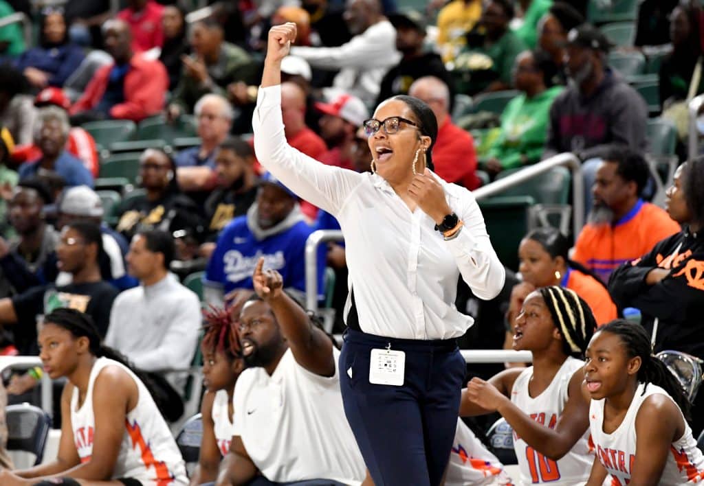 A basketball coach wearing a white blouse and dark pants stands on the sideline with her arm raised, shouting encouragement. She is surrounded by her team, who are sitting on the bench in white jerseys with orange accents. The background shows a crowd of spectators in various colorful attire.