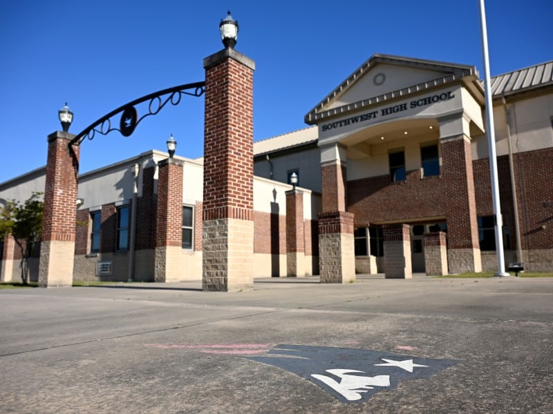 The image shows the front entrance of Southwest High School, featuring a building with red brick and beige stone accents. An archway with lamps on brick pillars stands before the entrance. The school name is visible on the building's facade, and a painted logo on the ground is partially seen in the foreground. The sky is clear and blue.