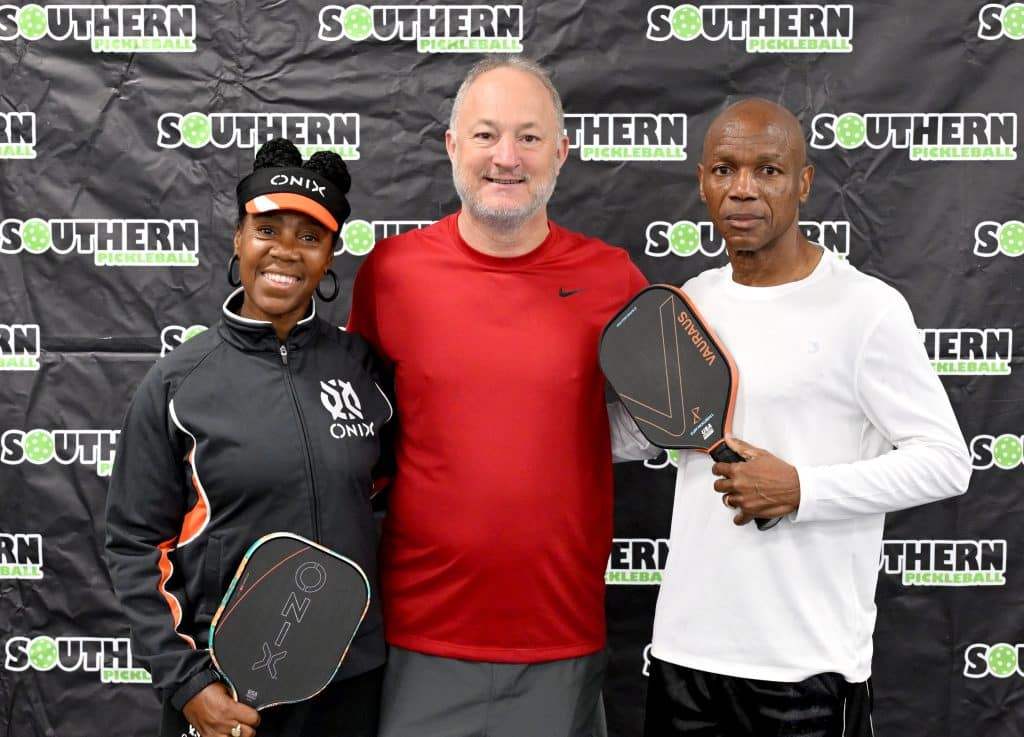 The mayor and the two tournament winners pose with pickleball paddles in front of a media backdrop.