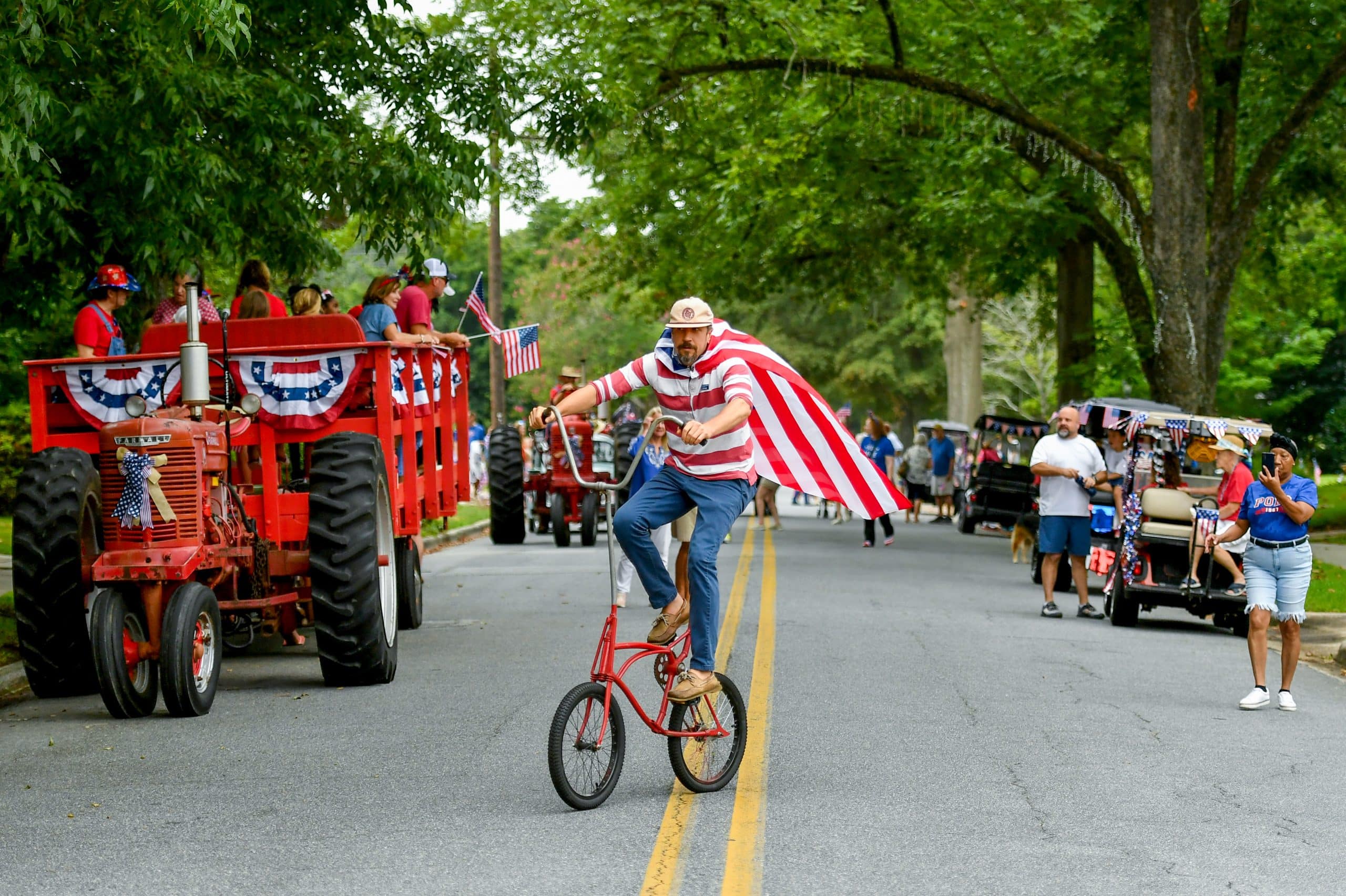 A man riding a bike and dressed in patriotic attire leads a neighborhood 4th of July parade.