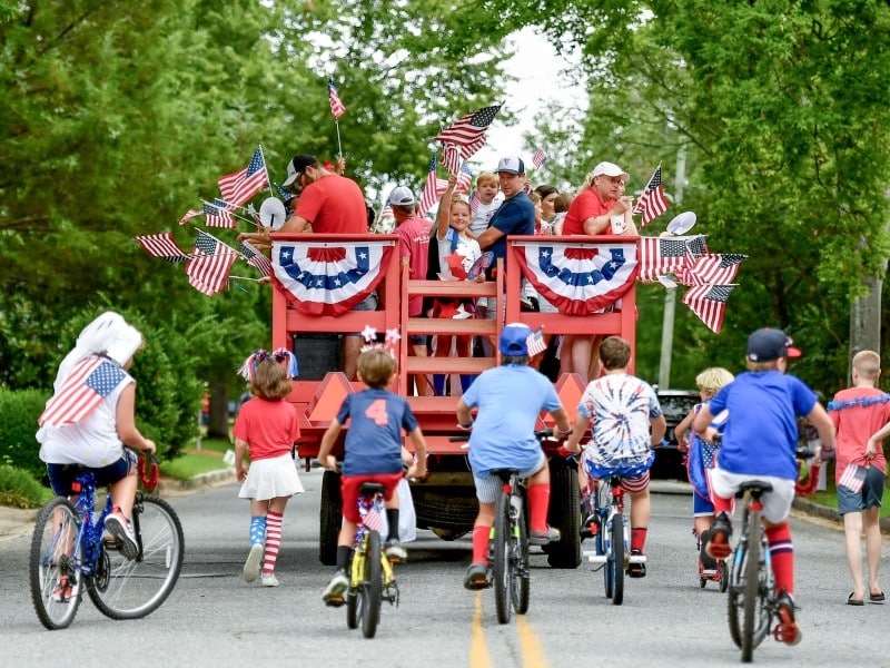 People wave from the back of a wagon while dressed in patriotic attire while riding down a street.
