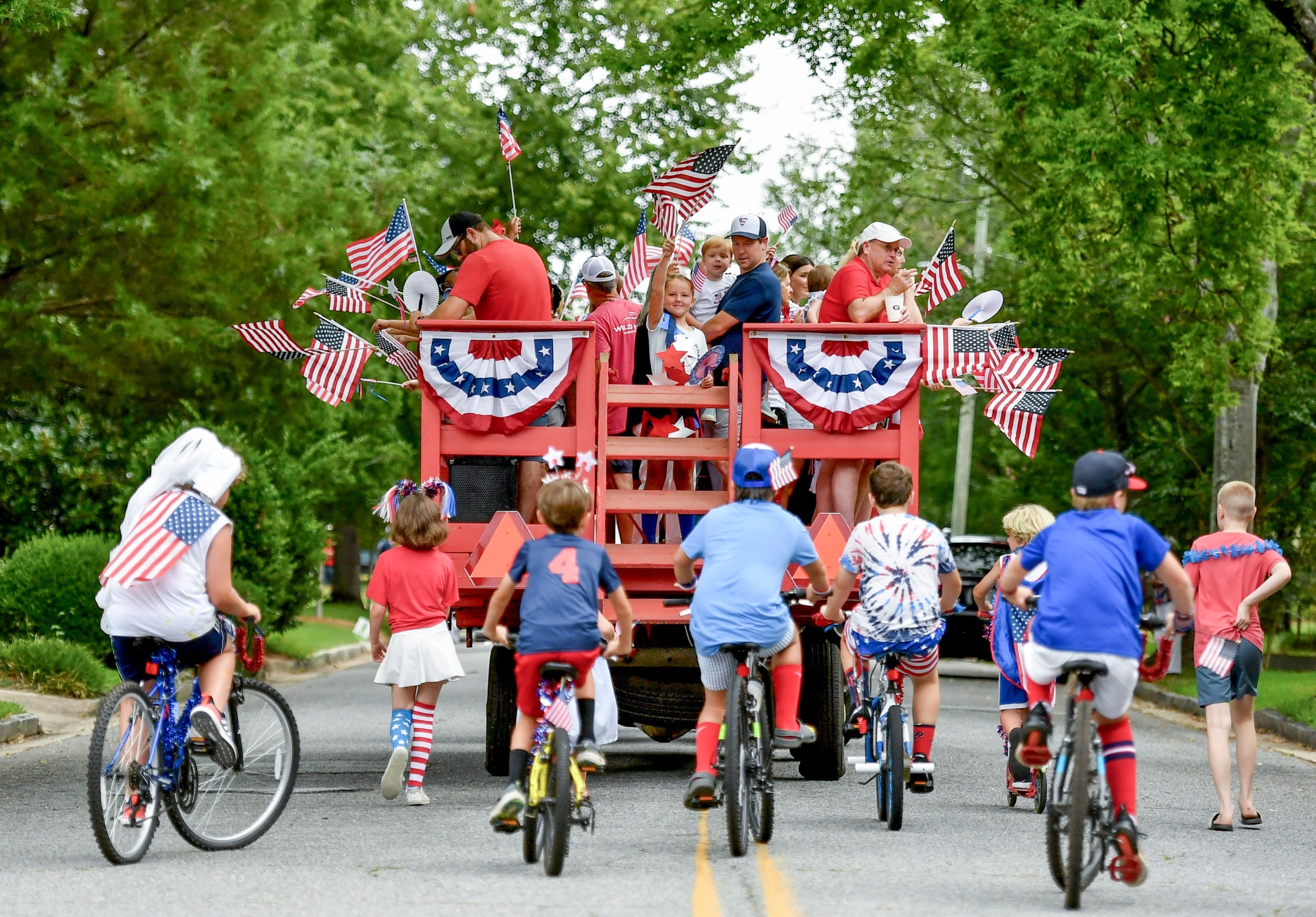 People wave from the back of a wagon while dressed in patriotic attire while riding down a street.