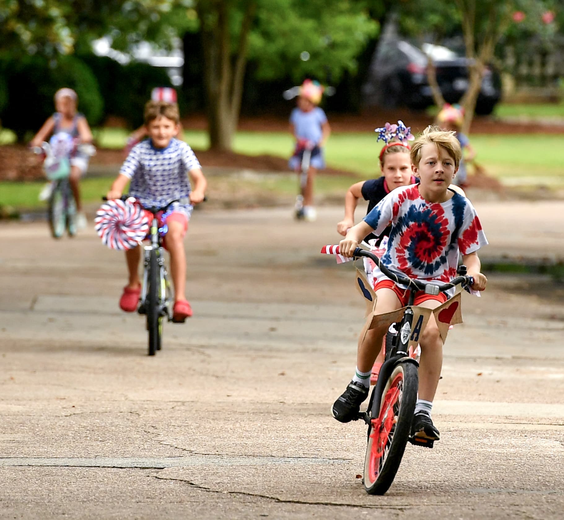 Children in patriotic attire ride bikes and scooters on the street.