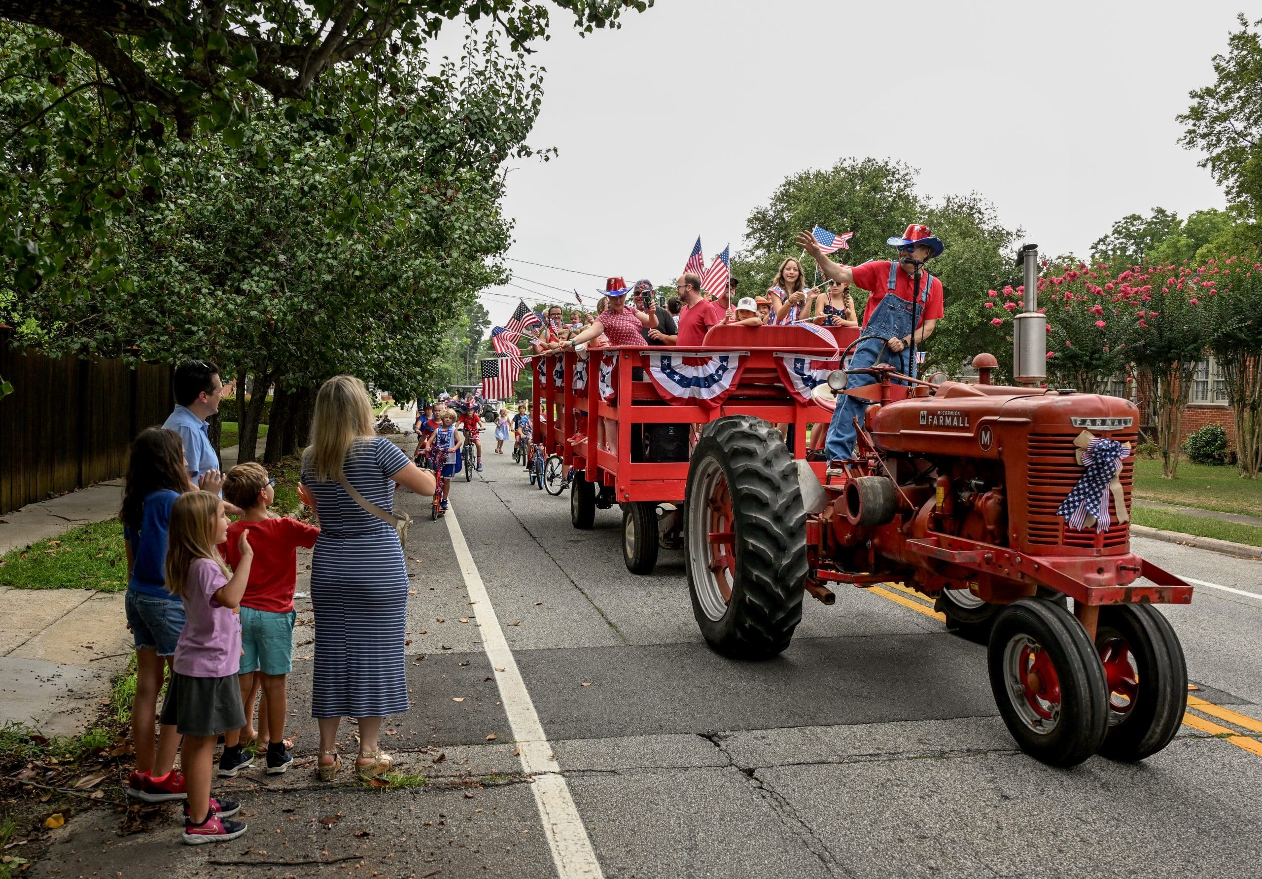 People wave as a tractor and wagon carrying people dressed in patriotic attire ride by along a street.