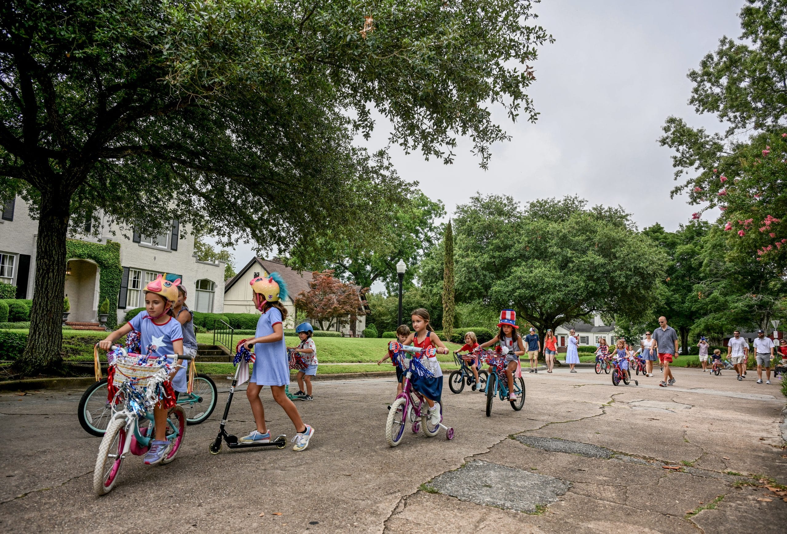 Children in patriotic attire ride bikes and scooters on the street.