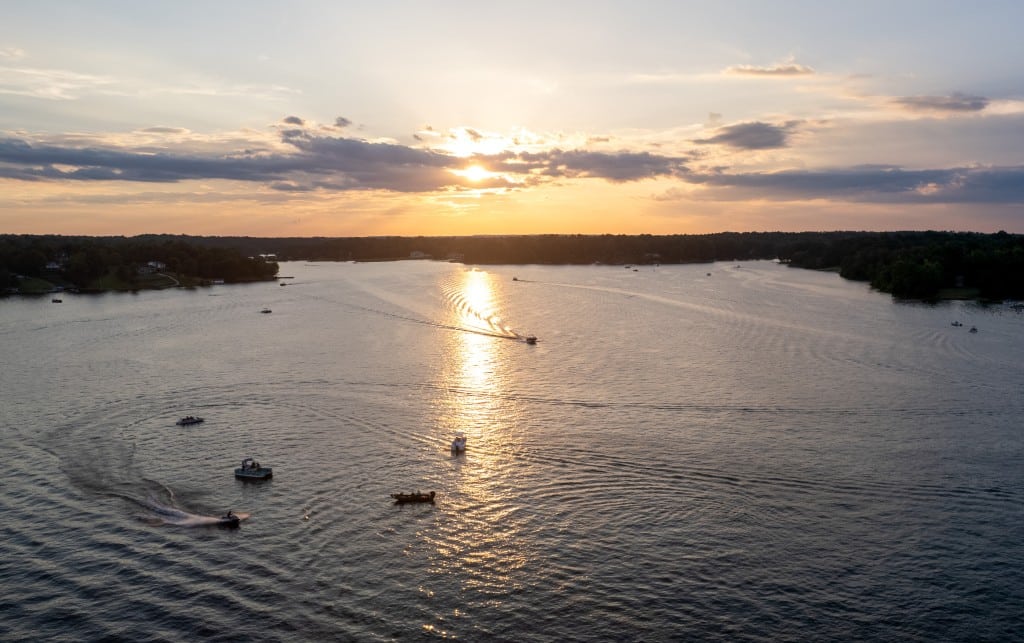 A sunset over a lake with several boats creating ripples on the water. The sun is low in the sky, casting a golden reflection on the lake's surface. The sky has a mix of clouds, and the shoreline is lined with trees and a few houses. The scene is calm and serene.