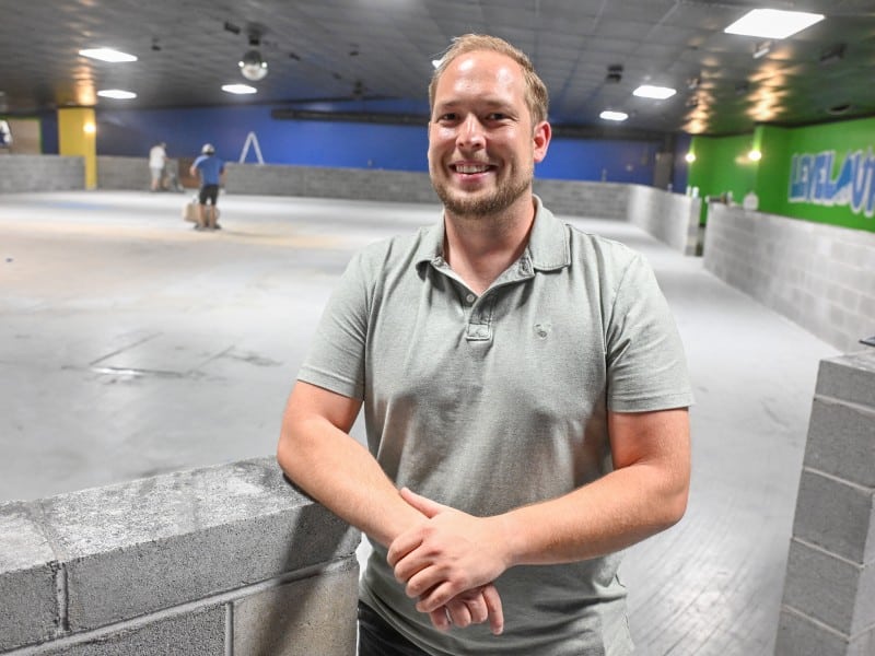 A man with a beard and short hair, wearing a light gray polo shirt, is standing and smiling in an indoor space under construction. In the background, there are several people working and cinder block walls with "LEVEL UP" painted in blue on a green section of the wall.