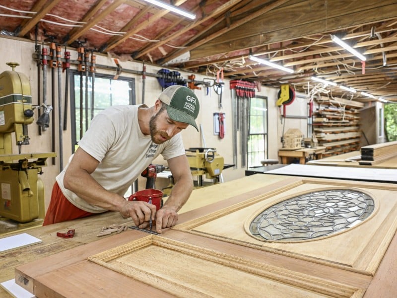 A man works on a door in his workshop, marking a spot with a pencil.