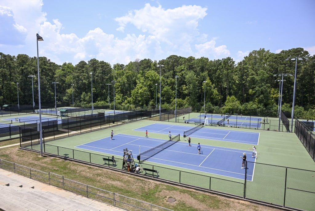 A wide photograph of blue tennis courts with green concrete surrounding them, all bordered in by fences and grass.