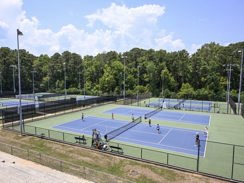 A wide photograph of blue tennis courts with green concrete surrounding them, all bordered in by fences and grass.