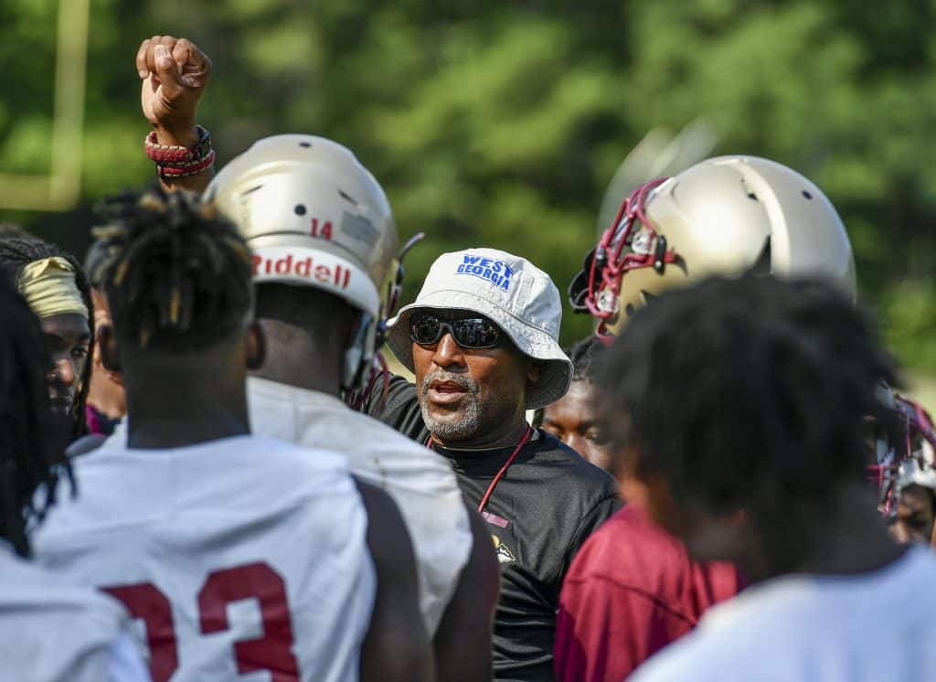 A football coach in a white bucket hat and black t-shirt raises his fist in the middle of a circle of players dressed in jerseys and helmets with no pads.
