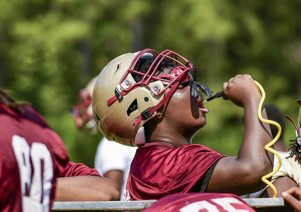 A football player dressed in a golden helmet and crimson jersey brings a yellow water hose to his mouth to take a drink while sitting on the bench at practice.