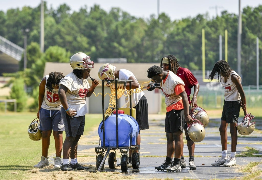 Several football players dressed in t-shirts, shorts and helmets with no pads hunch over to drink from spigots on a large water tank.