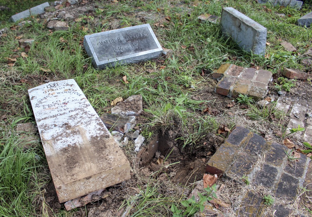 An old, damaged grave site with broken headstones and scattered bricks. One gravestone lies flat on the ground, partially covered with dirt. The area is overgrown with grass and fallen leaves.