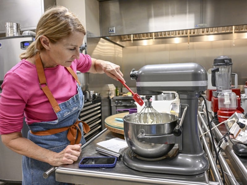 A woman with a red rubber spatula scrapes down the sides of a bowl under a stand mixer filled with butter and shortening.