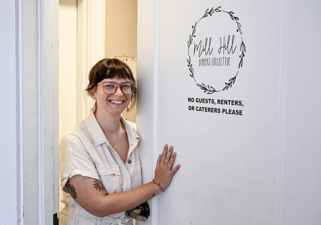 A woman stands next to a white door with the Mill Hill Bakers Collective logo painted on it.