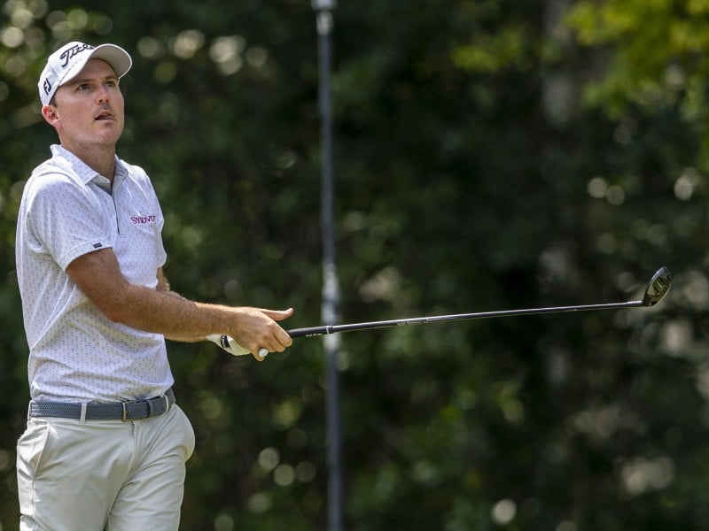 A golfer dressed in a white hat, patterned white polo and khaki pants holds his club to the right as he watches a shot travel.