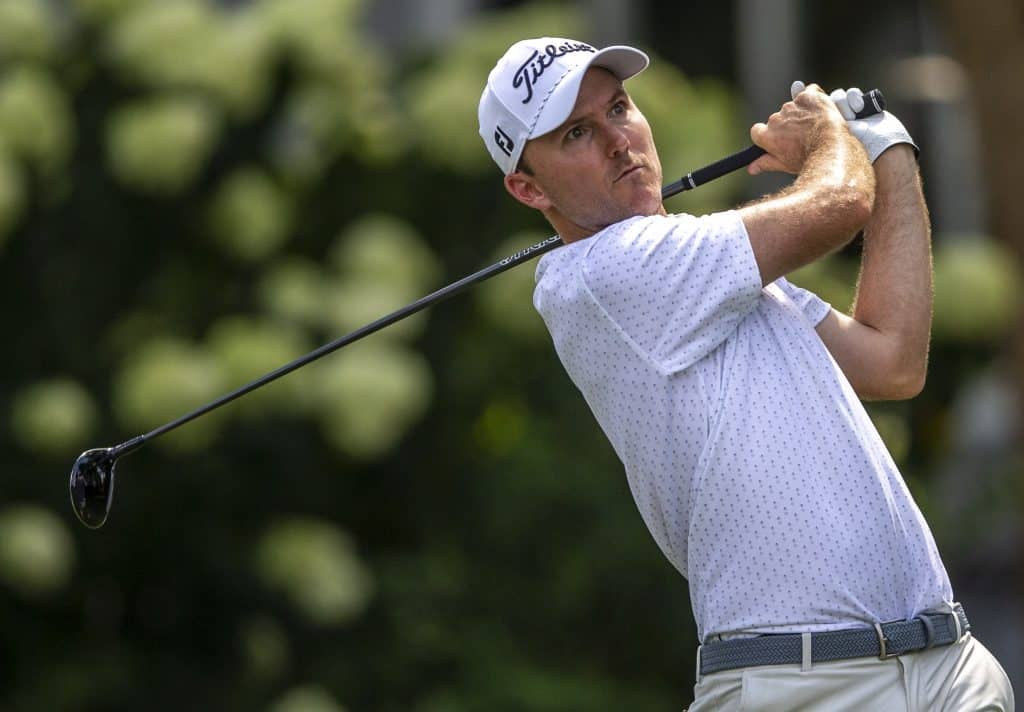 A man in a white hat and white patterned polo swings a golf club while gazing at his shot.