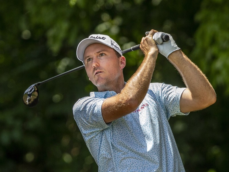 A golfer with a flower patterned shirt, white hat and glove watches his ball mid-stroke.