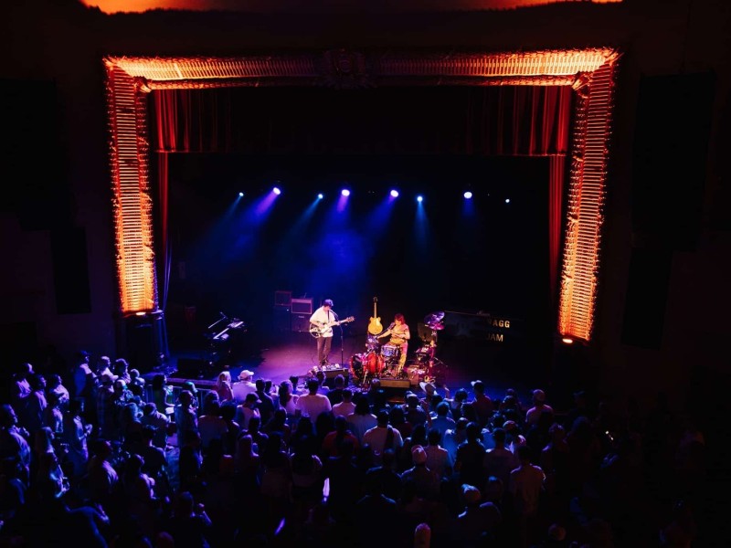A band performs on a stage lit with blue and purple lighting, framed by ornate red and orange theater curtains. The audience stands close to the stage, watching the performance. The scene is intimate, with a warm and vibrant atmosphere.