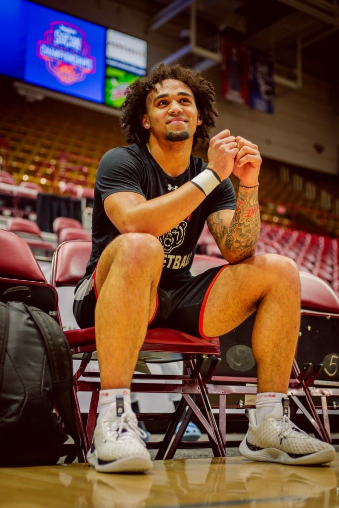 A basketball player in an all-black uniform with orange trim sits on the bench at a basketball court with his hands on his knees.