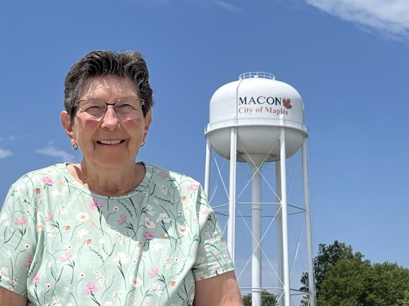 An older woman with short gray hair and glasses is smiling in front of a tall white water tower. The water tower has the text "MACON City of Maples" with a red maple leaf. The woman is wearing a light green shirt with a floral pattern. The sky is clear and blue with a few small clouds.