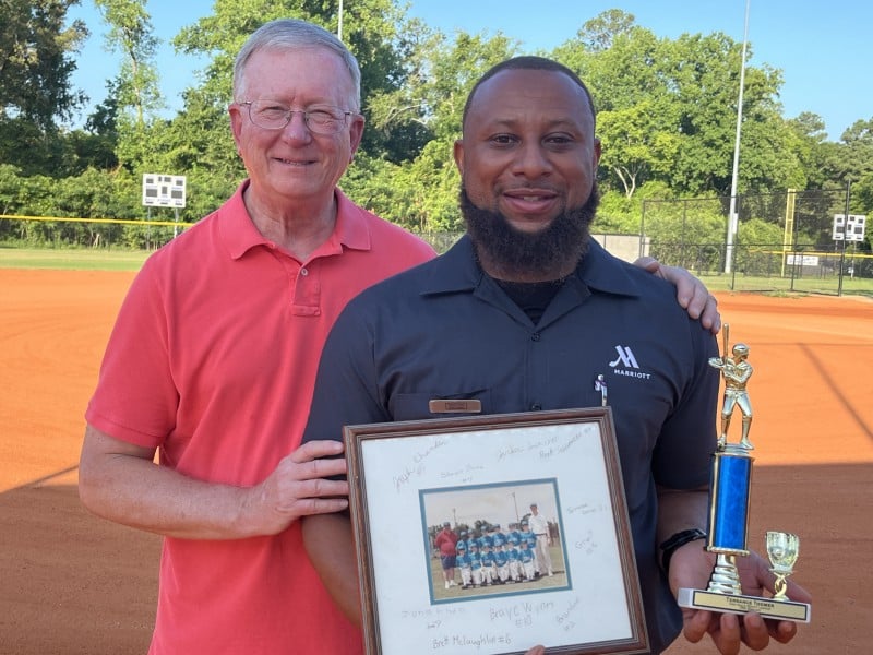 Two men stand on a baseball field, smiling at the camera. The older man on the left is wearing a red polo shirt and glasses, and has his hand on the shoulder of the younger man beside him. The younger man is wearing a black shirt with the "Marriott" logo and is holding a trophy and a framed photo of a baseball team. Trees and part of the baseball field are visible in the background.