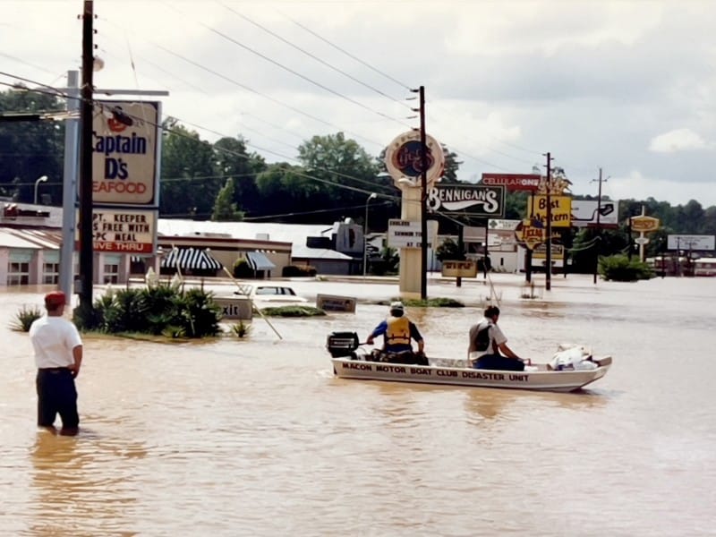 A photo shows a flooded city street, with two men in a boat and a third standing in the flood waters.