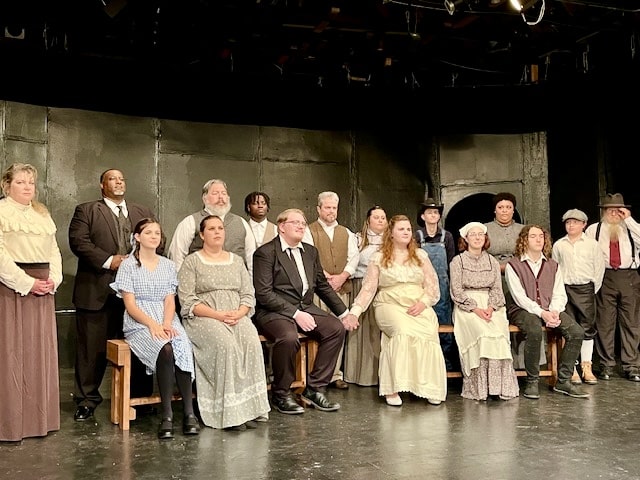 A group of fifteen people in period costumes poses on stage. Some are sitting on wooden benches while others stand behind them. The background is a dark, plain set. The people wear outfits ranging from formal suits to simple dresses and aprons, suggesting a historical setting.