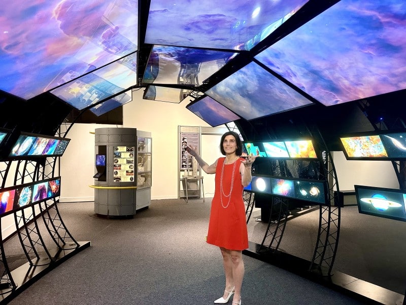 A woman in a red dress stands in an exhibit room with multiple curved screens displaying colorful space images. The ceiling features large monitors showing a nebula, while smaller screens around the room show various celestial objects. The woman is smiling with her arms slightly raised.