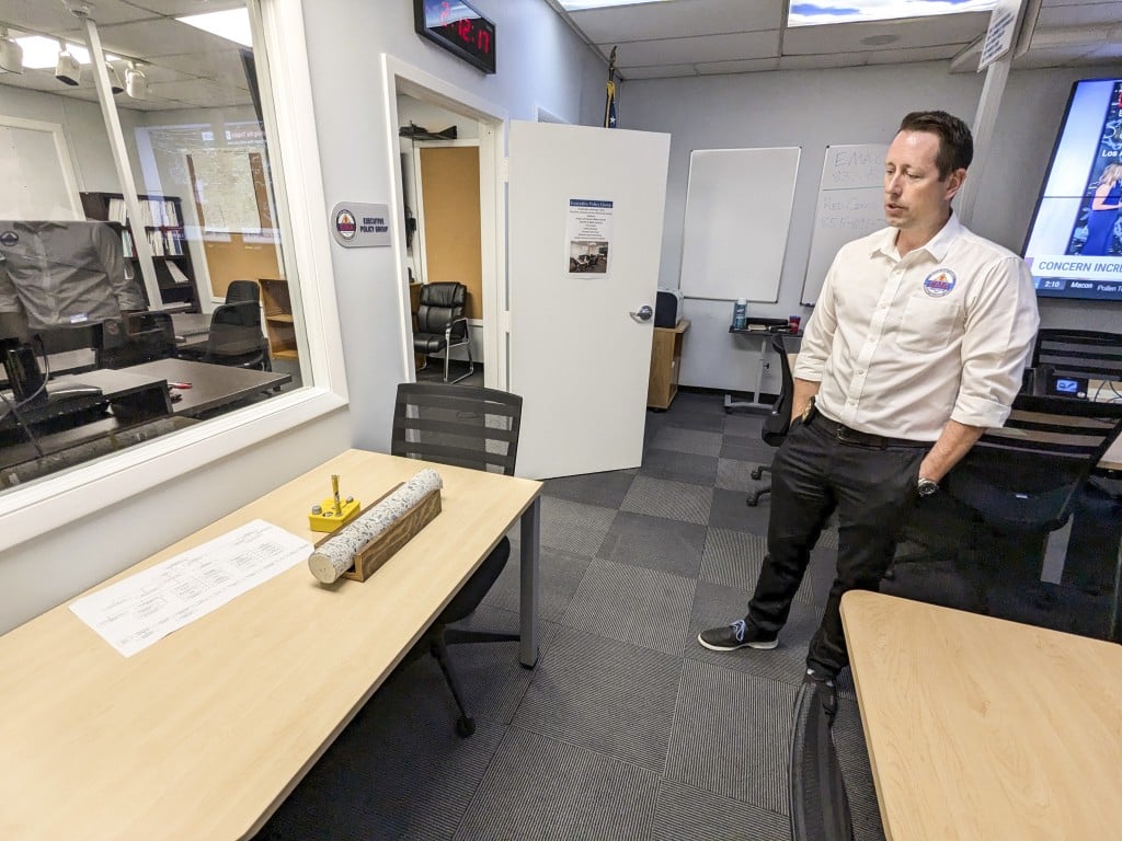 Stephen Hawkins, wearing a white shirt with an EMA logo stands in an office with multiple desks and chairs. He is looking at a cylindrical concrete log mounted on a wooden stand.