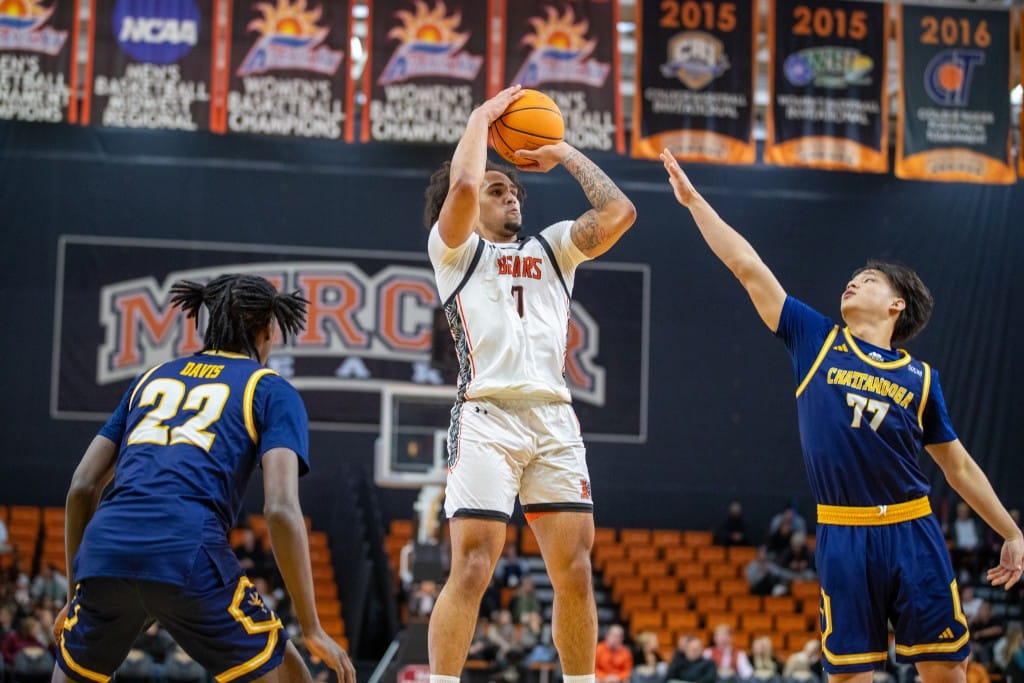 A basketball player in a white "Bears" jersey jumps to take a shot while two players in blue "Chattanooga" jerseys, one numbered 22 and the other 77, attempt to block him. The game takes place in a stadium with banners hanging above, and a "Mercer Bears" sign in the background.