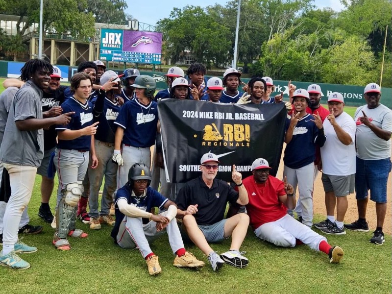 A baseball team celebrates on the field holding a black banner that reads "2024 Nike RBI Jr. Baseball Regionals Southern Region Champions." The players and coaches, wearing navy blue and white uniforms and hats, pose together, smiling and making celebratory gestures. A scoreboard and greenery are visible in the background.