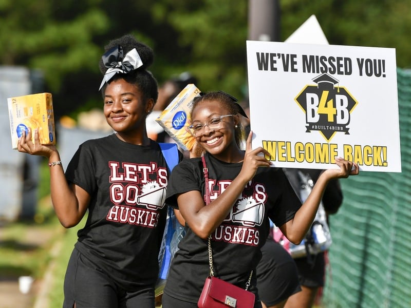 Two smiling young women are holding boxes of Dial soap and a welcome sign. They are wearing black T-shirts that read "LET'S GO HUSKIES" with a megaphone graphic. The sign one of them holds says, "WE'VE MISSED YOU! WELCOME BACK!" with the hashtag #BUILT4BIBB and the logo "B4B" on it. The background is blurred with green foliage and a chain-link fence.