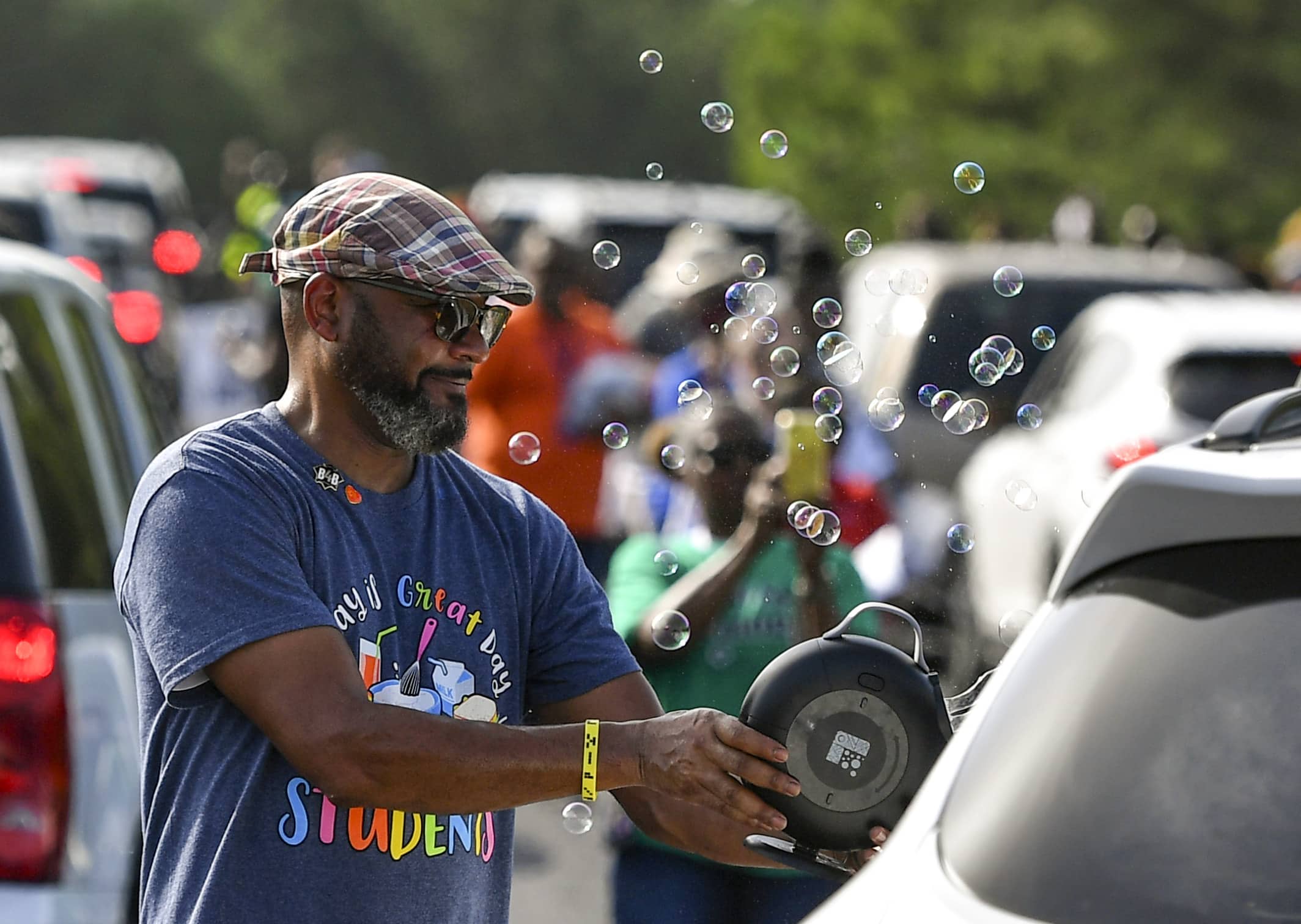 A man walks between a long long of automobiles while aiming a bubble machine t