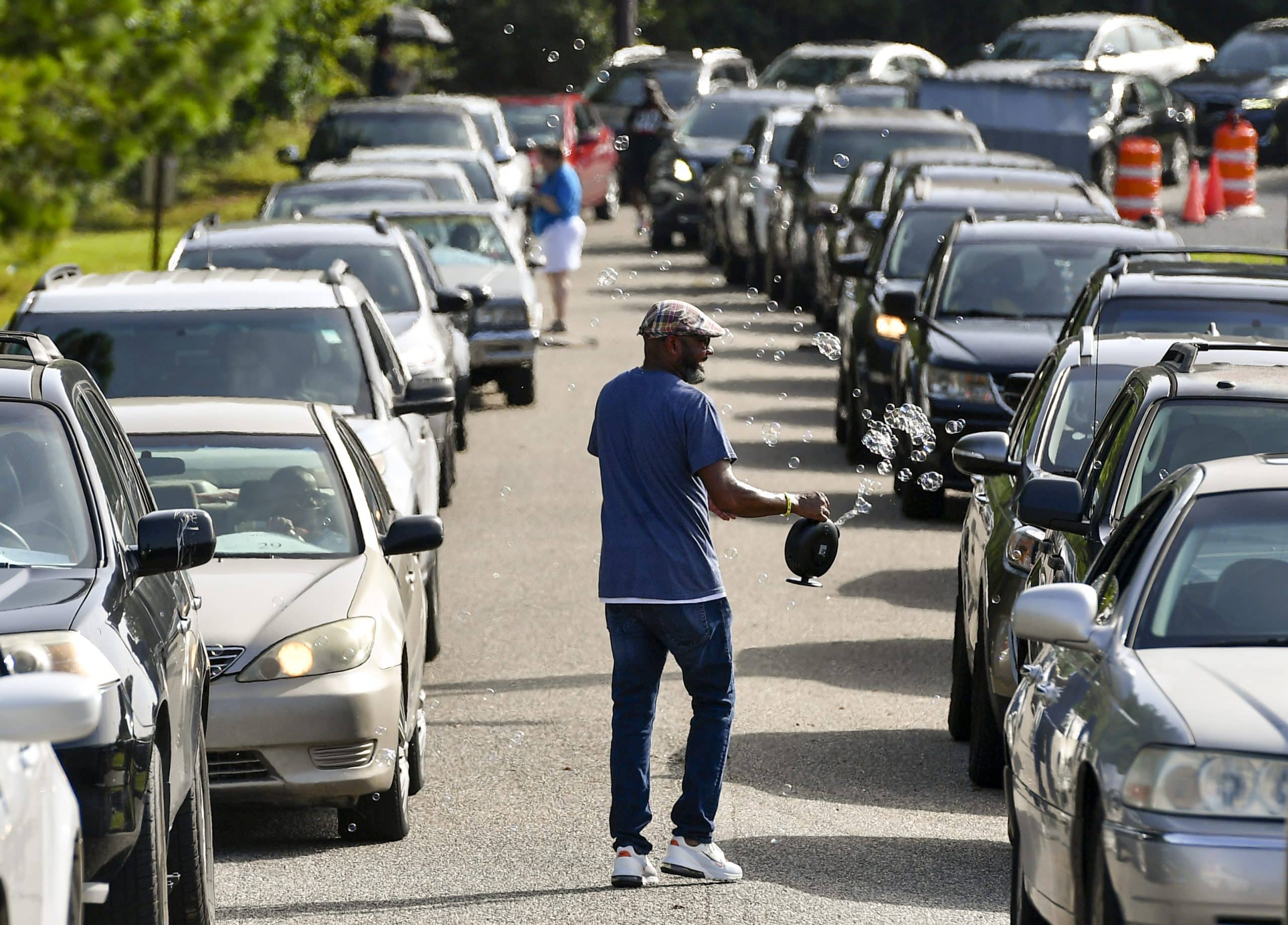 A man walks between a long long of automobiles while aiming a bubble machine towards a car.