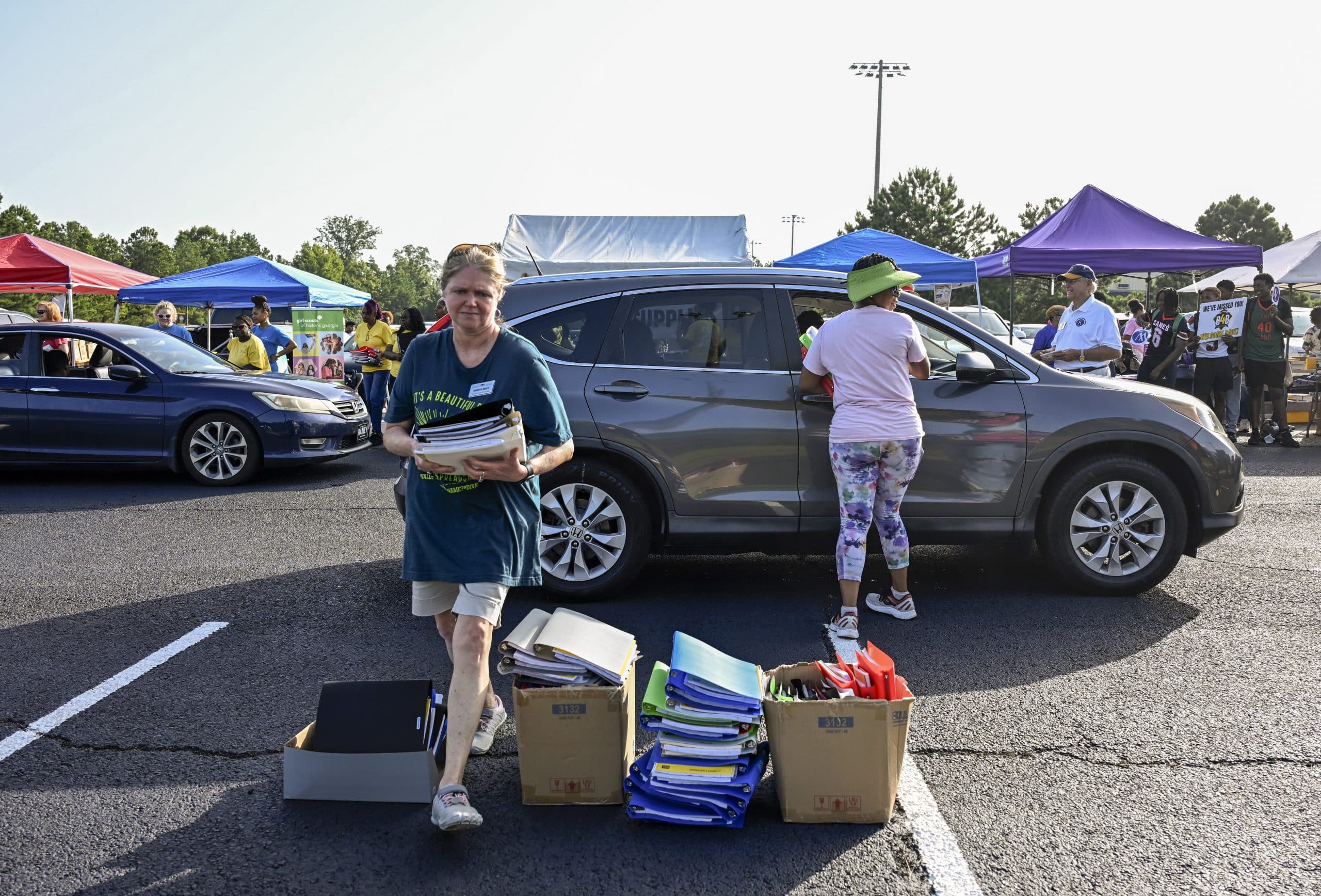 A woman hands school supplies through an SUV window, and another woman collects school supplies in the foreground.