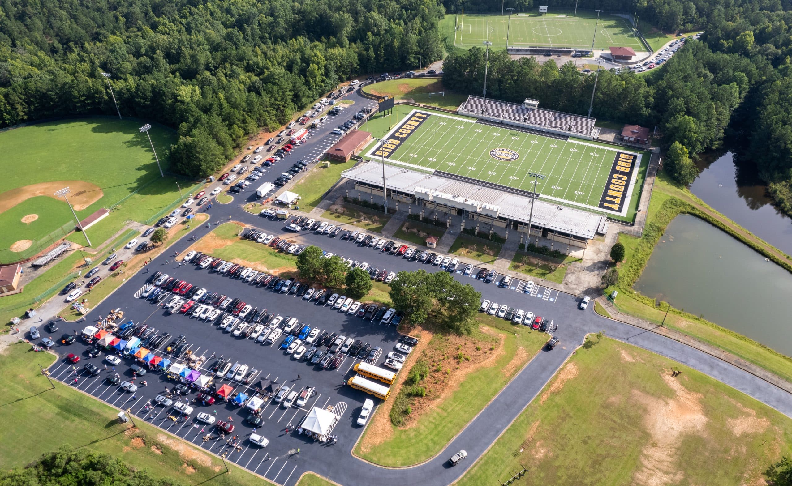 An aerial view of a sports complex surrounded by dense green forest. The complex includes a football stadium with "Bibb County" written on the field and a track surrounding it. Adjacent to the stadium are additional fields for soccer and baseball. The parking lot is filled with numerous cars, including a line of vehicles with colorful tents beside them, and a pair of yellow school buses parked nearby. A small pond and tree-lined pathways are visible on the right side of the image.