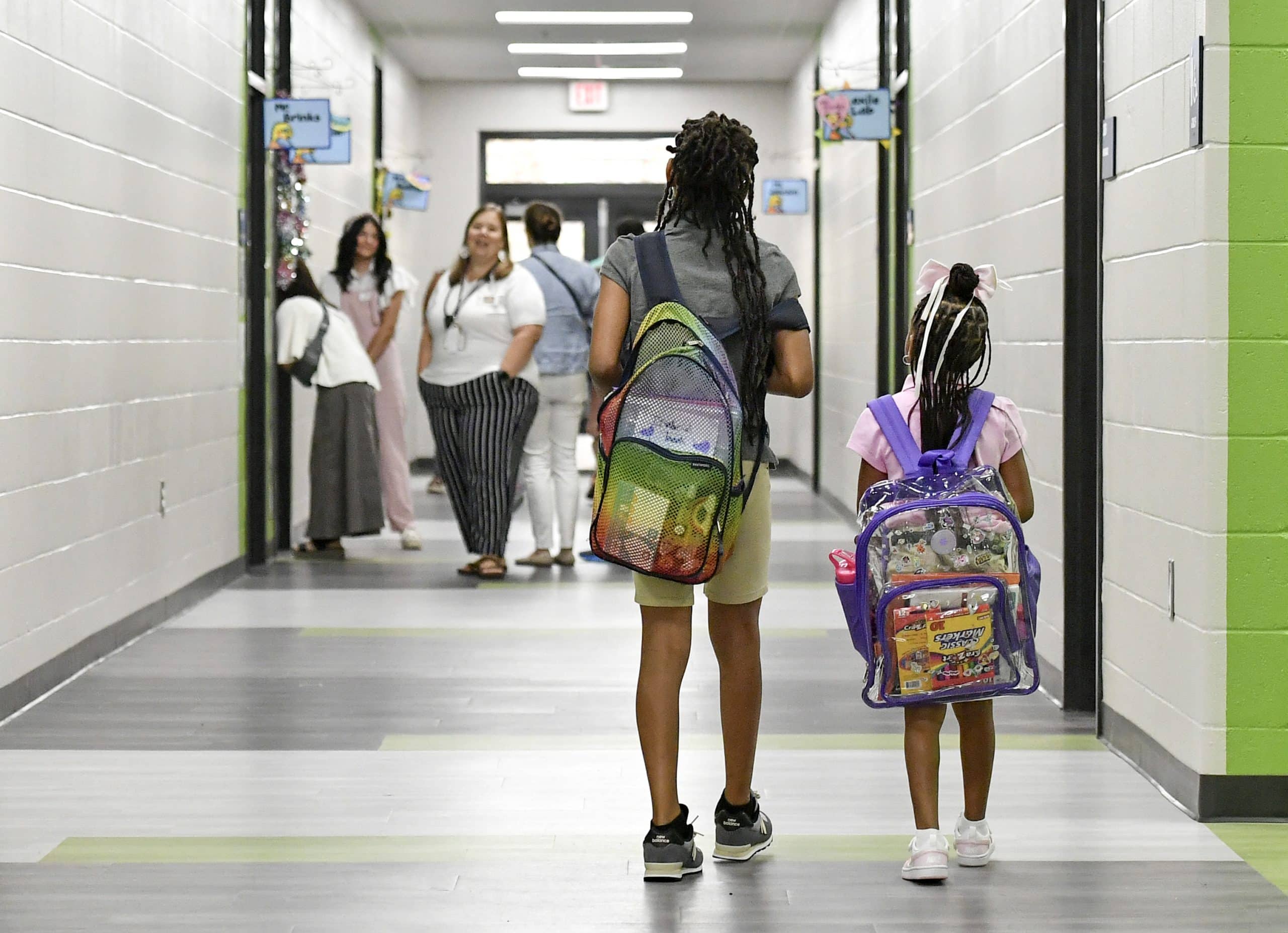 A photo from behind of two young children wearing backpacks walking down a school hallway.