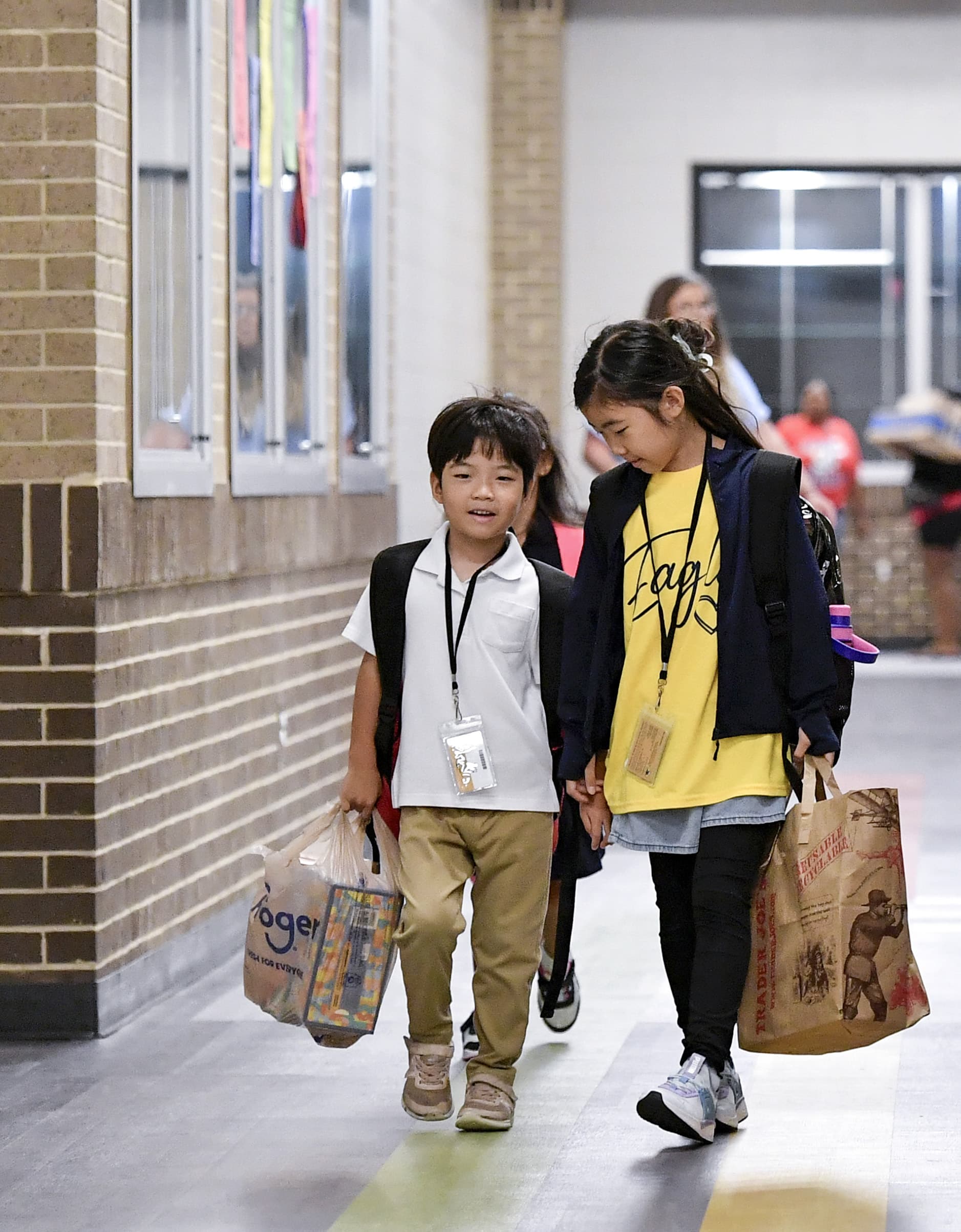 A photo of two young children wearing backpacks and carrying bags walking down a school hallway.
