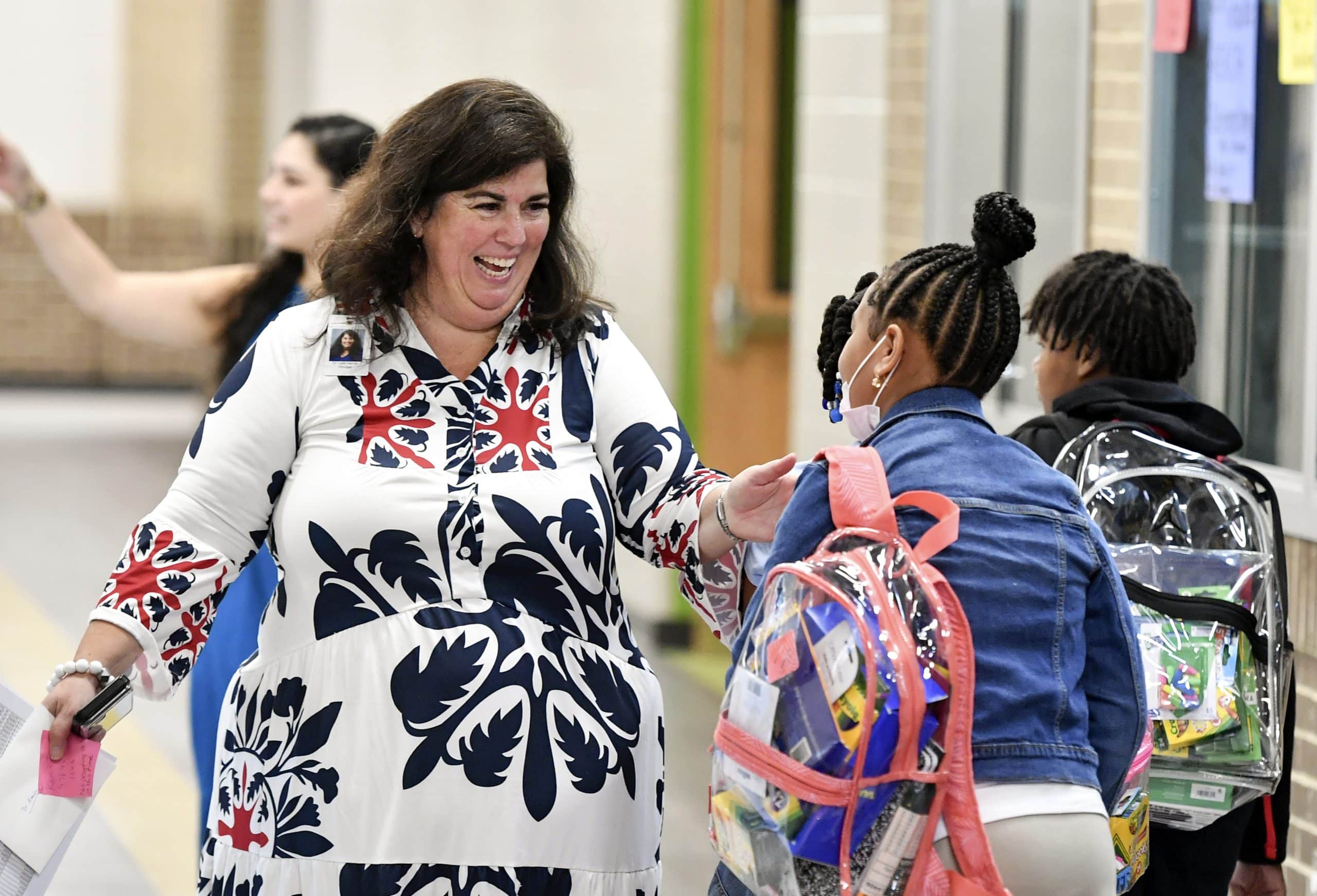 A woman opens her arms to receive a hug from a child wearing a backpack in a school hallway.